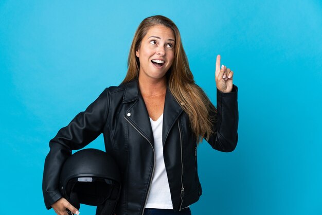 Vrouw van middelbare leeftijd die een motorhelm houdt die op blauwe muur wordt geïsoleerd en een idee denkt dat de vinger benadrukt