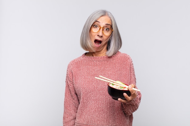 Vrouw van middelbare leeftijd die erg geschokt of verrast kijkt, starend met open mond en zegt wow Aziatisch voedselconcept