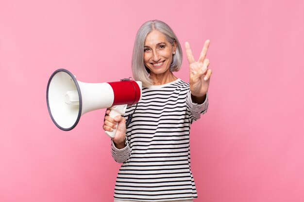 Vrouw van middelbare leeftijd die lacht en er gelukkig, zorgeloos en positief uitziet, gebarend overwinning of vrede met één hand met een megafoon