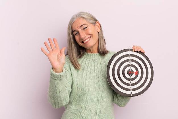 Vrouw van middelbare leeftijd die lacht en vrolijk zwaait met de hand die je verwelkomt en je darts-doelconcept begroet