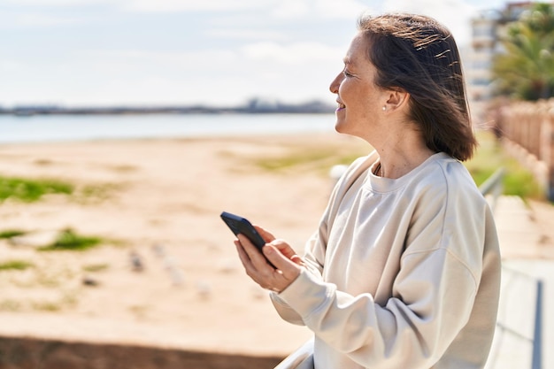 Vrouw van middelbare leeftijd die lacht zelfverzekerd met behulp van smartphone aan zee