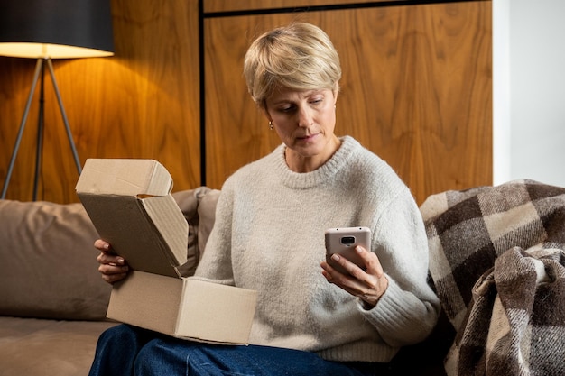 Vrouw van middelbare leeftijd die op de bank zit, opent de doos en controleert het pakket met een smartphone