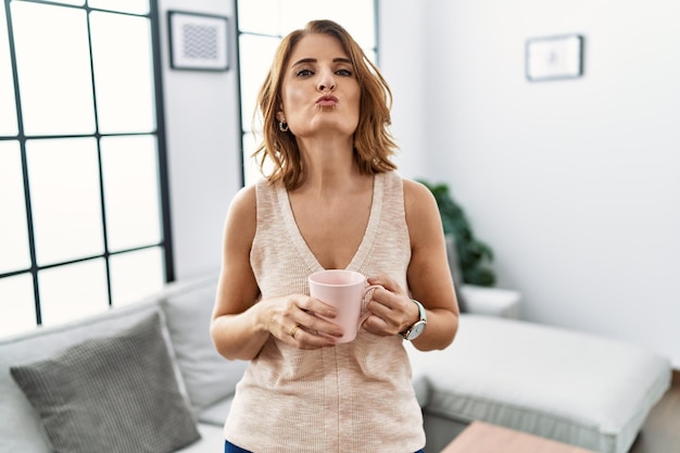 Vrouw van middelbare leeftijd die thuis een kopje koffie drinkt en naar de camera kijkt terwijl ze een kus in de lucht blaast en een mooie en sexy liefdesuitdrukking is