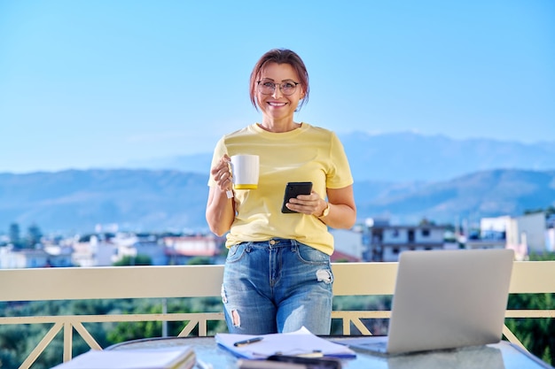 Vrouw van middelbare leeftijd die thuis werkt op terras ontspannend met mok thee en smartphone