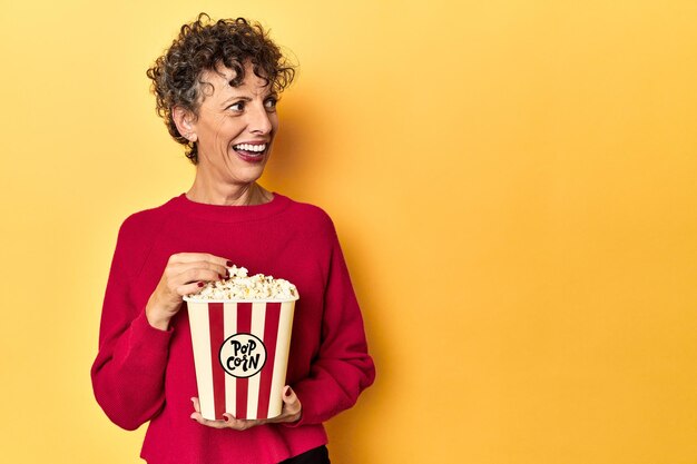 Vrouw van middelbare leeftijd die van popcorn geniet voor een film op een gele studioachtergrond