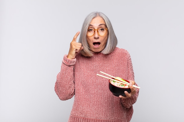 Vrouw van middelbare leeftijd die verrast, met open mond, geschokt kijkt, een nieuwe gedachte, idee of concept Aziatisch voedselconcept realiseert
