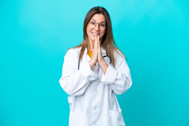 Vrouw van middelbare leeftijd geïsoleerd op een blauwe achtergrond, gekleed in een doktersjas en smekend