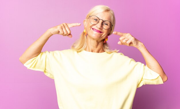 Vrouw van middelbare leeftijd glimlachend vol vertrouwen wijzend op een eigen brede glimlach, positieve, ontspannen, tevreden houding