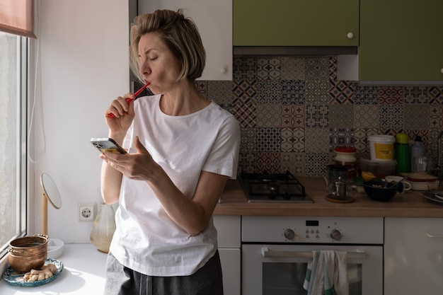 Vrouw van middelbare leeftijd in de ochtend thuis tanden poetsen en mobiele telefoon gebruiken
