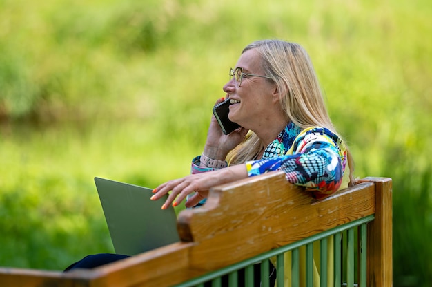 Vrouw van middelbare leeftijd in de tuin die vanuit huis werkt met behulp van laptop en aan de telefoon spreekt