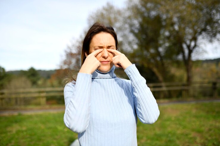 Vrouw van middelbare leeftijd in het park of in het veld die in haar ogen wrijft vanwege allergie of stof die in de herfst of winter een coltrui draagt