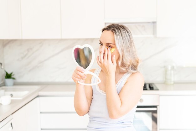 Vrouw van middelbare leeftijd in pyjama in de keuken met cosmetische collageenpleisters onder de ogen Gezichtsverzorging Huidcosmetica Ochtendroutine Thuis spa en wellness cosmetische ingrepen Ooghuidbehandeling