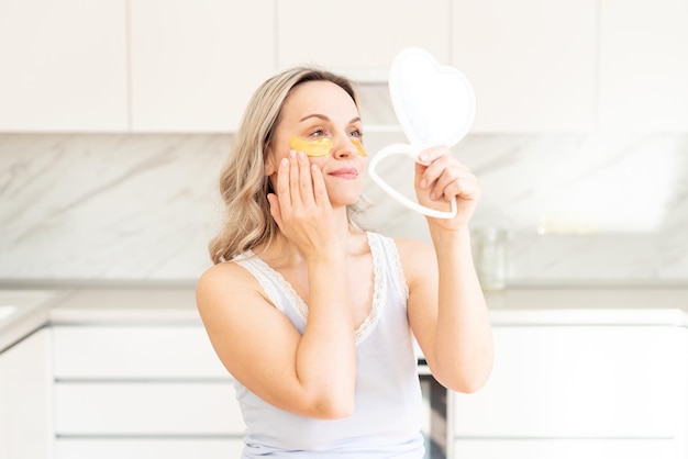 Vrouw van middelbare leeftijd in pyjama in de keuken met cosmetische collageenpleisters onder de ogen Gezichtsverzorging Huidcosmetica Ochtendroutine Thuis spa en wellness cosmetische ingrepen Ooghuidbehandeling