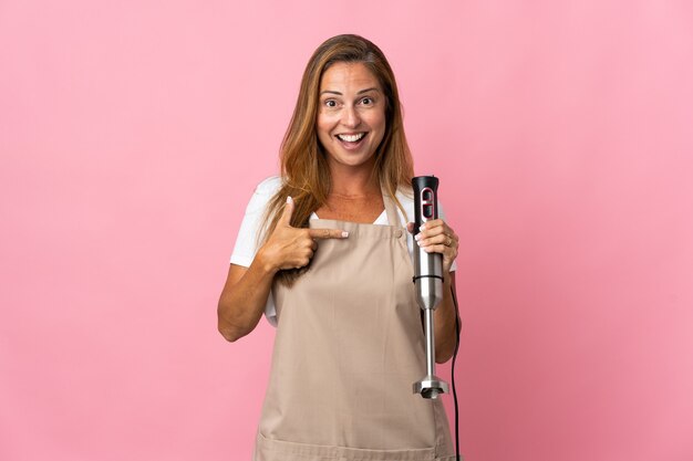 Vrouw van middelbare leeftijd met behulp van staafmixer geïsoleerd op roze met verrassingsgelaatsuitdrukking