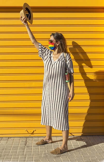 Vrouw van middelbare leeftijd met een hoed, een zonnebril en een regenboogkleurig beschermend masker. LGTB op gele achtergrond. Covid-19 concept