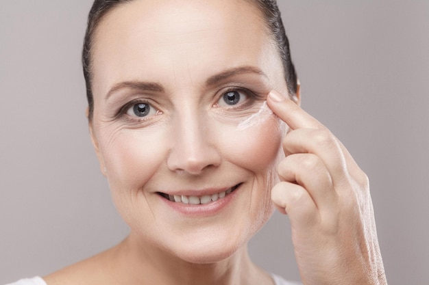 Vrouw van middelbare leeftijd met een perfecte gezichtshuid die cosmetische crème op de huid in de buurt van de ogen aanbrengt en naar de camera kijkt met een brede glimlach geïsoleerd op een grijze achtergrond. Huidverzorgingsconcept, kopieerruimte, studio-opname, binnen