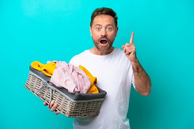 Vrouw van middelbare leeftijd met een wasmand geïsoleerd op een blauwe achtergrond denkend aan een idee dat met de vinger omhoog wijst