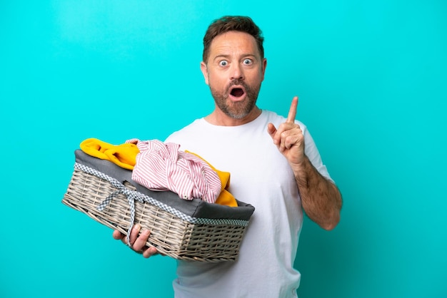 Vrouw van middelbare leeftijd met een wasmand geïsoleerd op een blauwe achtergrond die de oplossing wil realiseren terwijl ze een vinger opheft