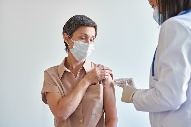 Vrouw van middelbare leeftijd met gezichtsmasker die op het kantoor van de dokter zit en covid-opname krijgt tijdens pandemie