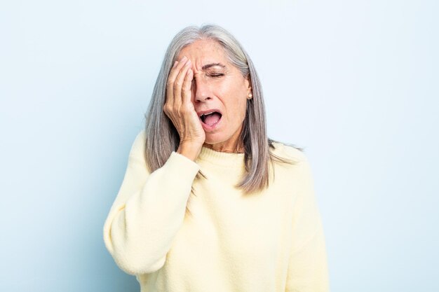 Vrouw van middelbare leeftijd met grijs haar die er slaperig, verveeld en geeuwend uitziet, met hoofdpijn en een hand die het halve gezicht bedekt