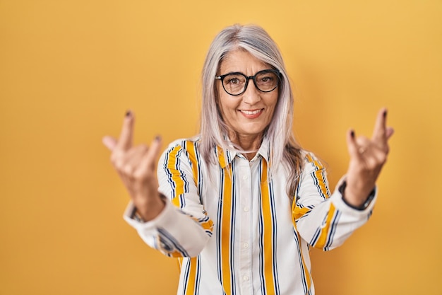 Foto vrouw van middelbare leeftijd met grijs haar die over een gele achtergrond staat en een bril draagt die schreeuwt met een gekke uitdrukking die een rocksymbool doet met de handen omhoog. muziek ster. zwaar concept.