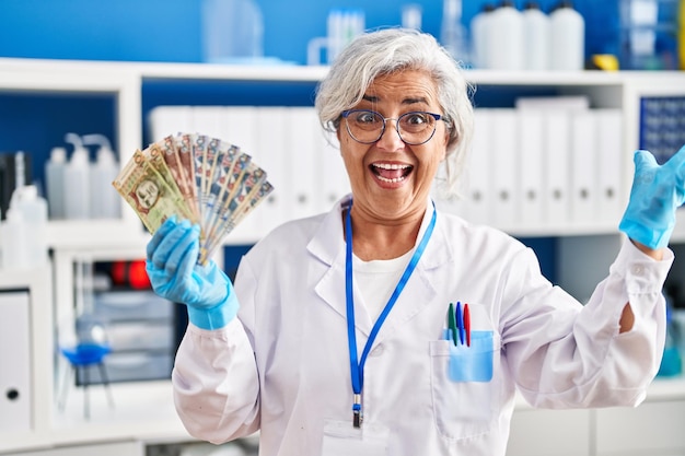 Vrouw van middelbare leeftijd met grijs haar die werkt in het laboratorium van een wetenschapper met Poolse zloty-bankbiljetten die de overwinning vieren met een gelukkige glimlach en uitdrukking van de winnaar met opgeheven handen