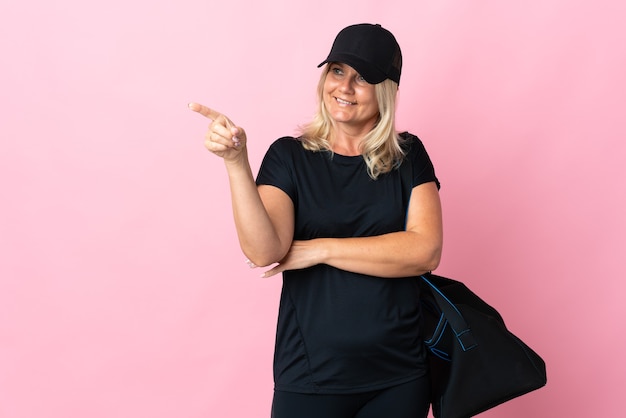 Vrouw van middelbare leeftijd met sporttas geïsoleerd op roze muur wijzende vinger naar de zijkant