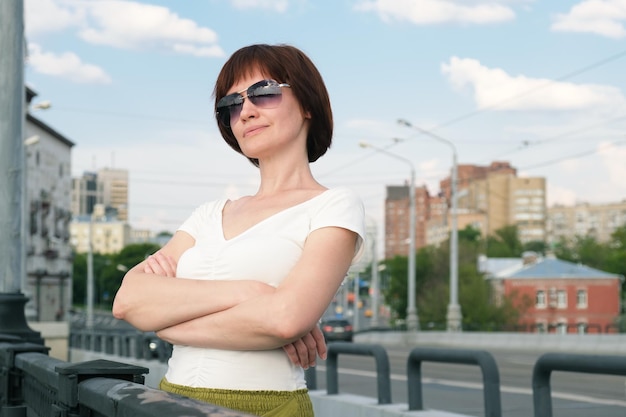 Vrouw van middelbare leeftijd met zonnebril staat met haar armen gekruist tegen de achtergrond van een stadsgezicht