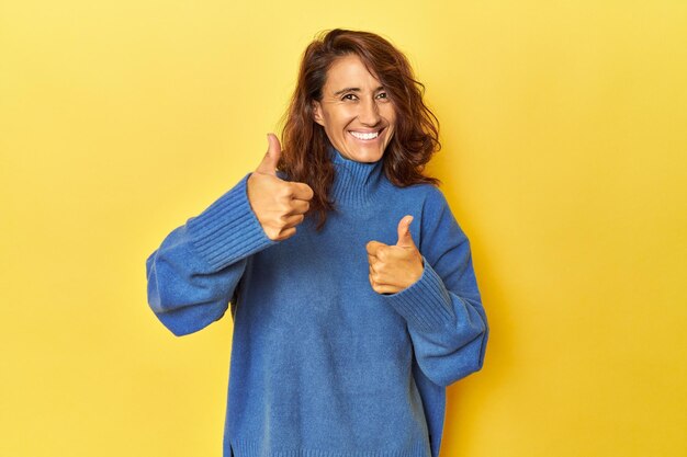 Foto vrouw van middelbare leeftijd op een gele achtergrond die beide duimen omhoog steekt, glimlachend en zelfverzekerd