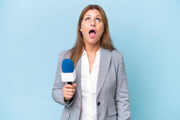 Foto vrouw van middelbare leeftijd tv-presentator over geïsoleerde blauwe achtergrond opkijkend en met een verbaasde uitdrukking