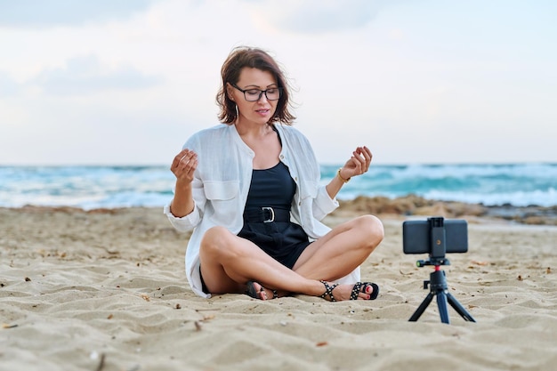 Vrouw van middelbare leeftijd zittend op het strand met smartphone via videogesprek