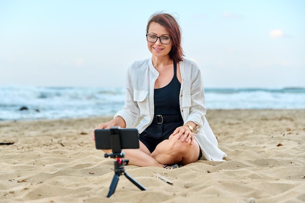 Vrouw van middelbare leeftijd zittend op het strand met smartphone via videogesprek