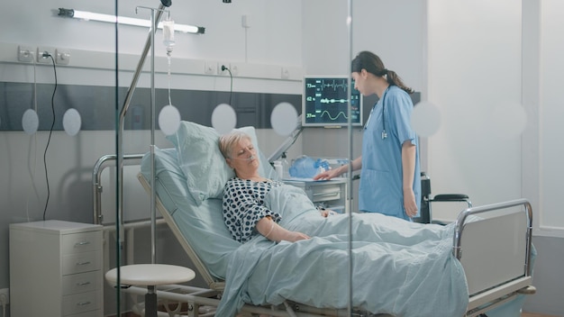 Vrouw verpleegster controleren op slapende oude patiënt in bed voor behandeling in de gezondheidszorg. Medisch assistent kijken naar hartslagmeter voor polsmeting. Specialist en gepensioneerde op ziekenhuisafdeling