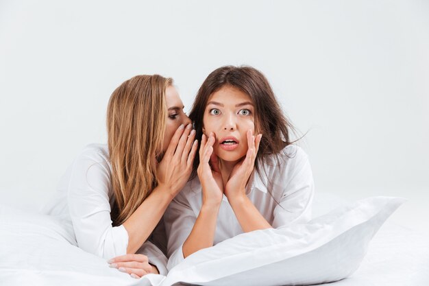 Vrouw vertelt roddels aan haar vriendin terwijl ze op bed ligt met kussens