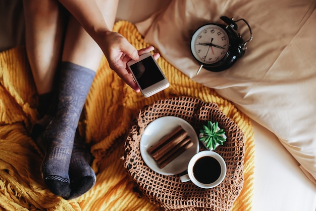 Foto vrouw voeten op sokken ontspannen op het bed met smartphone en koffie bij het ontbijt