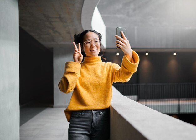 Vrouw vrede en selfie in kantoorgebouw glimlach en gelukkig terwijl op internet pose en emoji Aziatisch meisje en zakelijke ondernemer met smartphone voor foto vredesteken en hand voor profielfoto