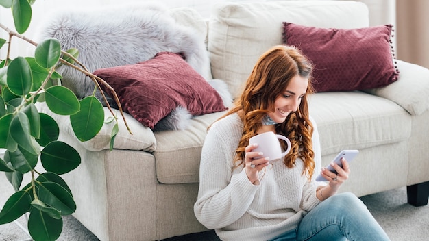 vrouw vrije tijd. gezellige huiselijke sfeer. inhoud jonge dame zittend op de vloer met kopje browsen telefoon.