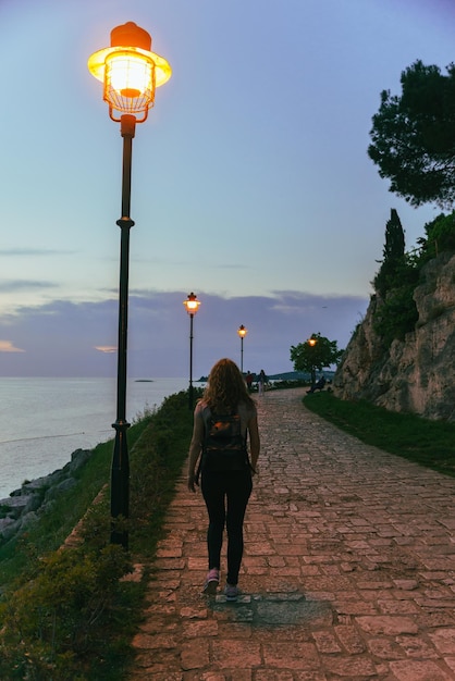 Vrouw wandelen door park in de buurt van zee op zonsondergang stadslicht lampen