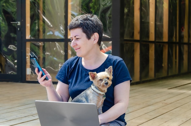 Vrouw werkt buiten op laptop, houdt Yorkshire Terrier-hond in haar armen en praat aan de telefoon