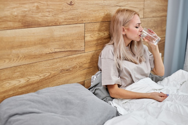 Vrouw zich ziek voelen, drinkwater met pillen medicijnen, interieur, kopie ruimte. Nog blonde dame met koorts, verkouden of coronavirus-infectie. alleen in de slaapkamer