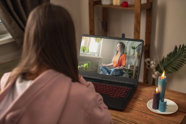 Vrouw zit aan een tafel voor een laptop te mediteren en te ontspannen terwijl ze online luistert naar een instructeur tijdens de videosessie Concept van een therapie op afstand en meditatie met een coach