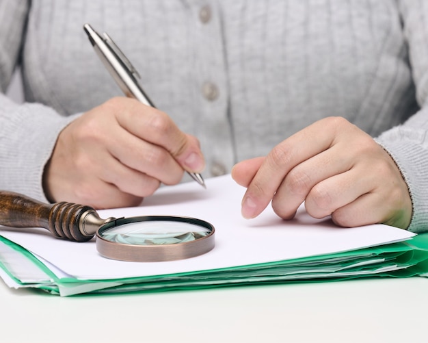 Vrouw zit aan een witte tafel en houdt een metalen pen boven een stapel papieren, in de andere hand een vergrootglas. Fouten vinden, het budget analyseren