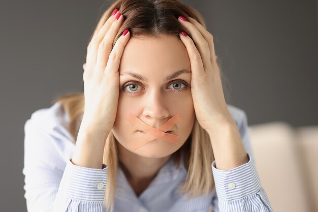 Foto vrouw zit aan tafel met haar mond verzegeld met gips en houdt haar hoofd vast. huiselijk geweld tegen vrouwen concept