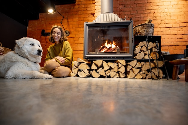 Vrouw zit bij de open haard met een witte hond in een gezellig en loft-achtig interieur