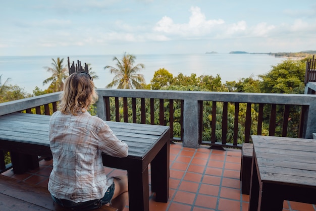 Vrouw zit bij de tafel op een leeg terras van café of huis en kijkt naar zeelandschap, palmbomen. Balkon op een tropisch eiland in Thailand