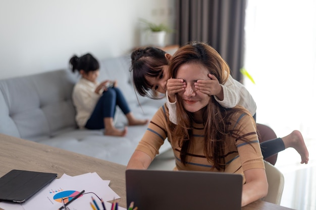 Vrouw zit bij de tafel op kantoor aan huis tijdens lockdown, werken op laptop. Speels kind leidt af van het werk, bedekt de ogen van haar moeder, kind maakt lawaai en vraagt aandacht van drukke moeder