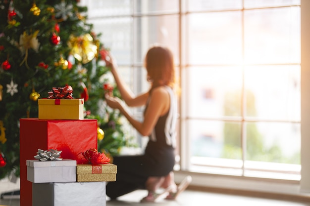 Vrouw zit en versier kerstbomen in het huis naast een stapel geschenkdozen met warm licht van het glazen raam aan de achterkant.