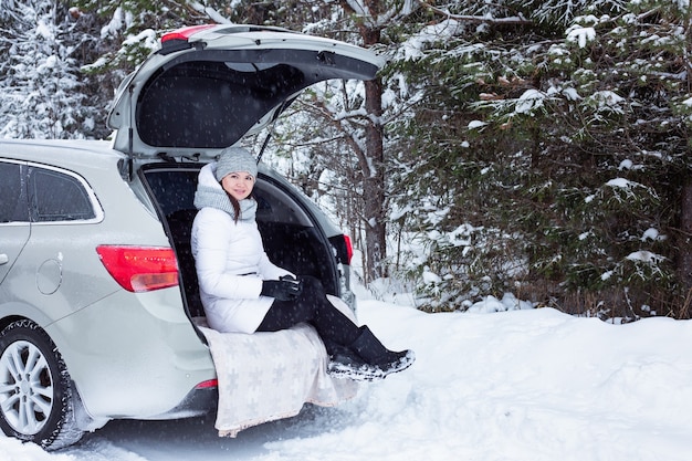 Vrouw zit in de kofferbak van de auto en houdt een kop hete thee in handen. Wintervakantie, reizen. Sneeuw bos en weg. Sneeuwweer - Afbeelding