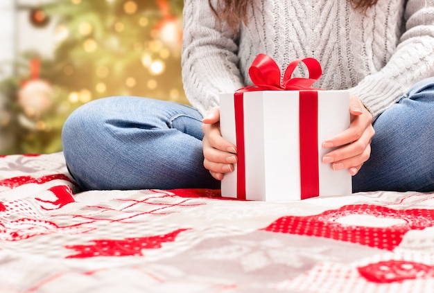 Vrouw zit met een cadeau in haar handen op het bed tegen de achtergrond van een kerstboom