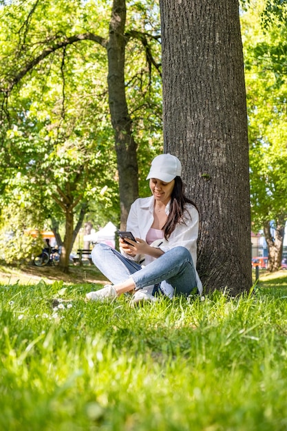 Vrouw zit onder de boom in het stadspark en surft op internet aan de telefoon